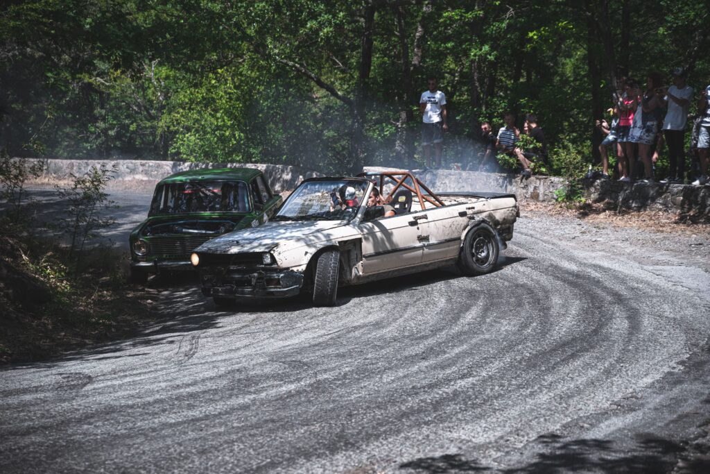 Exciting rally scene with cars drifting around a tight forest bend, watched by spectators.